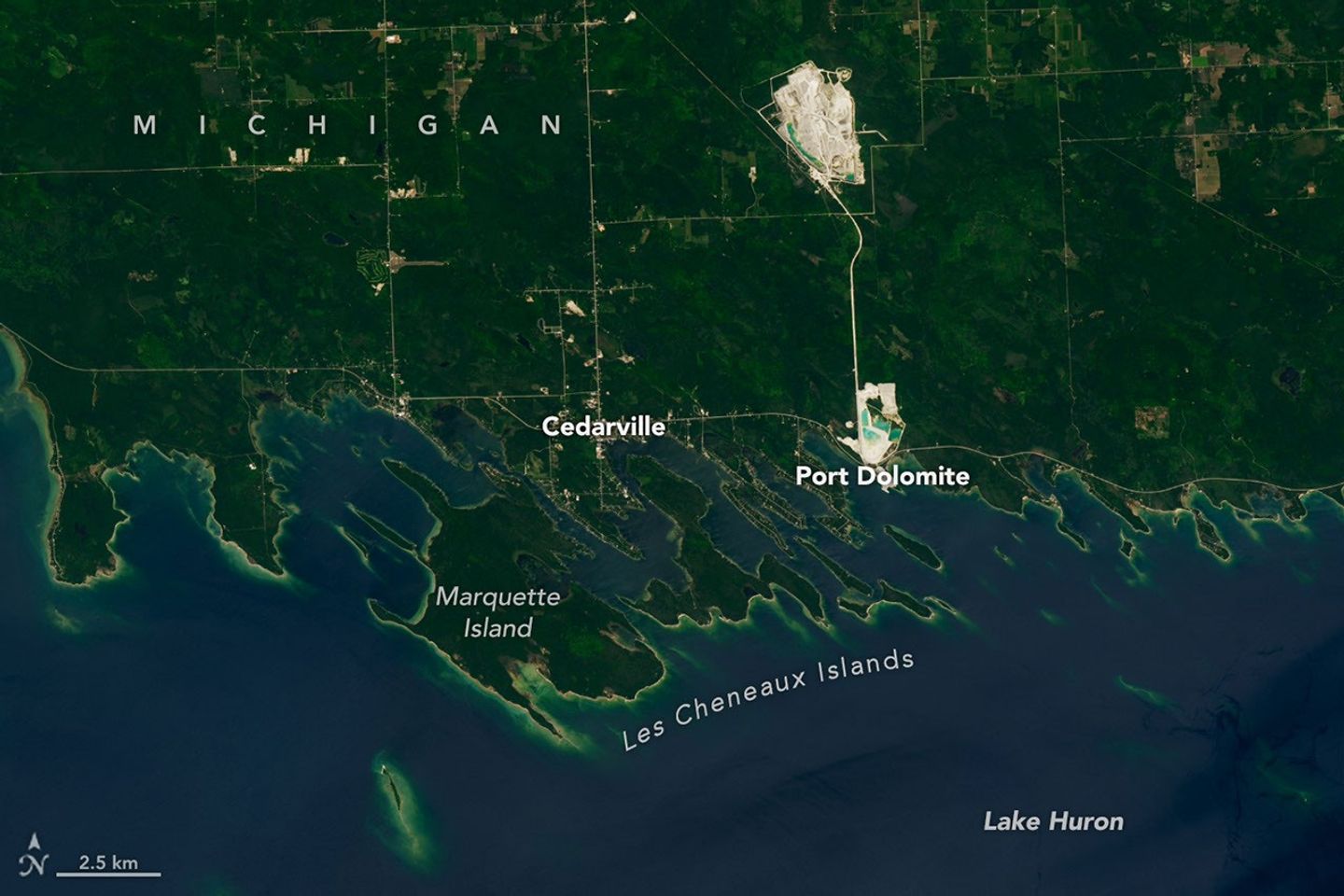 A portion of the Upper Peninsula of Michigan runs across the top of the image. It is mostly green, with some roads and two bright rock quarries visible. Numerous islands near the shore have elongated shapes and are oriented at a diagonal.