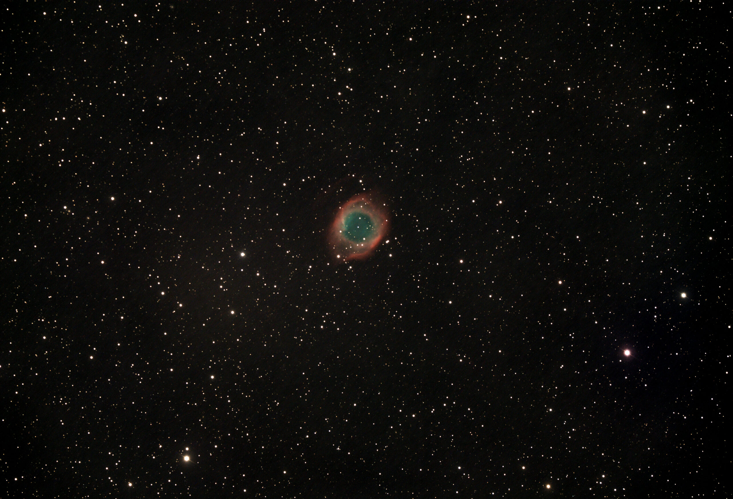 A telescopic photograph of the Helix Nebula (NGC 7293), a large planetary nebula appearing as a circular ring of glowing teal-green gas surrounded by a diffuse red outer halo, set against a black sky scattered with orange and white foreground stars.