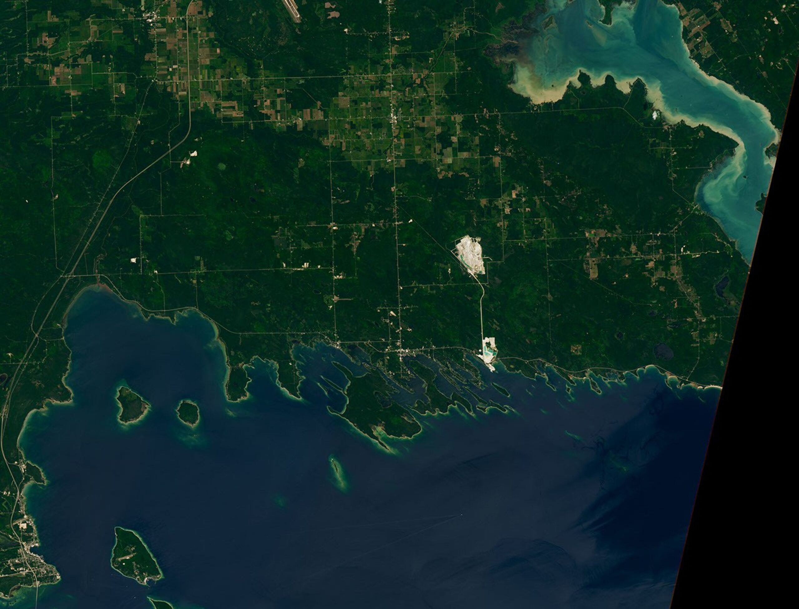 A portion of the Upper Peninsula of Michigan runs across the top of the image. It is mostly green, with some roads and two bright rock quarries visible. Numerous islands near the shore have elongated shapes and are oriented at a diagonal.
