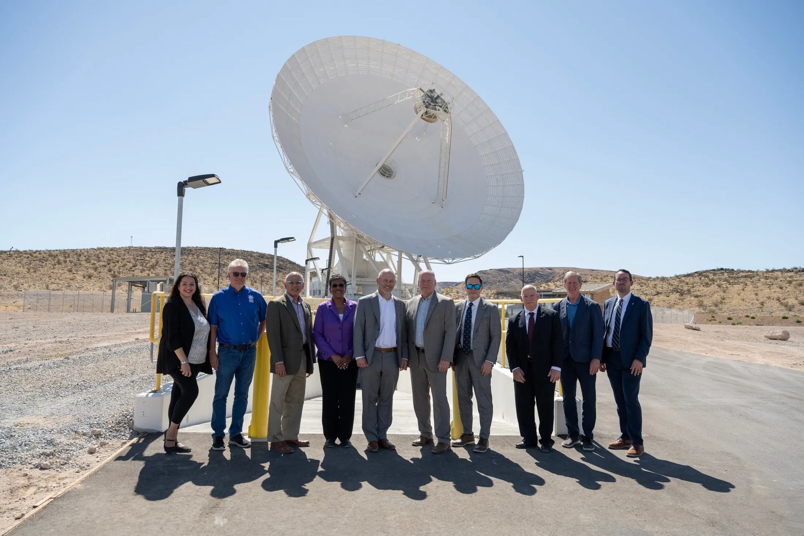 Ten people in professional attire pose together outside under a clear blue sky, with a massive white satellite dish standing directly behind them.