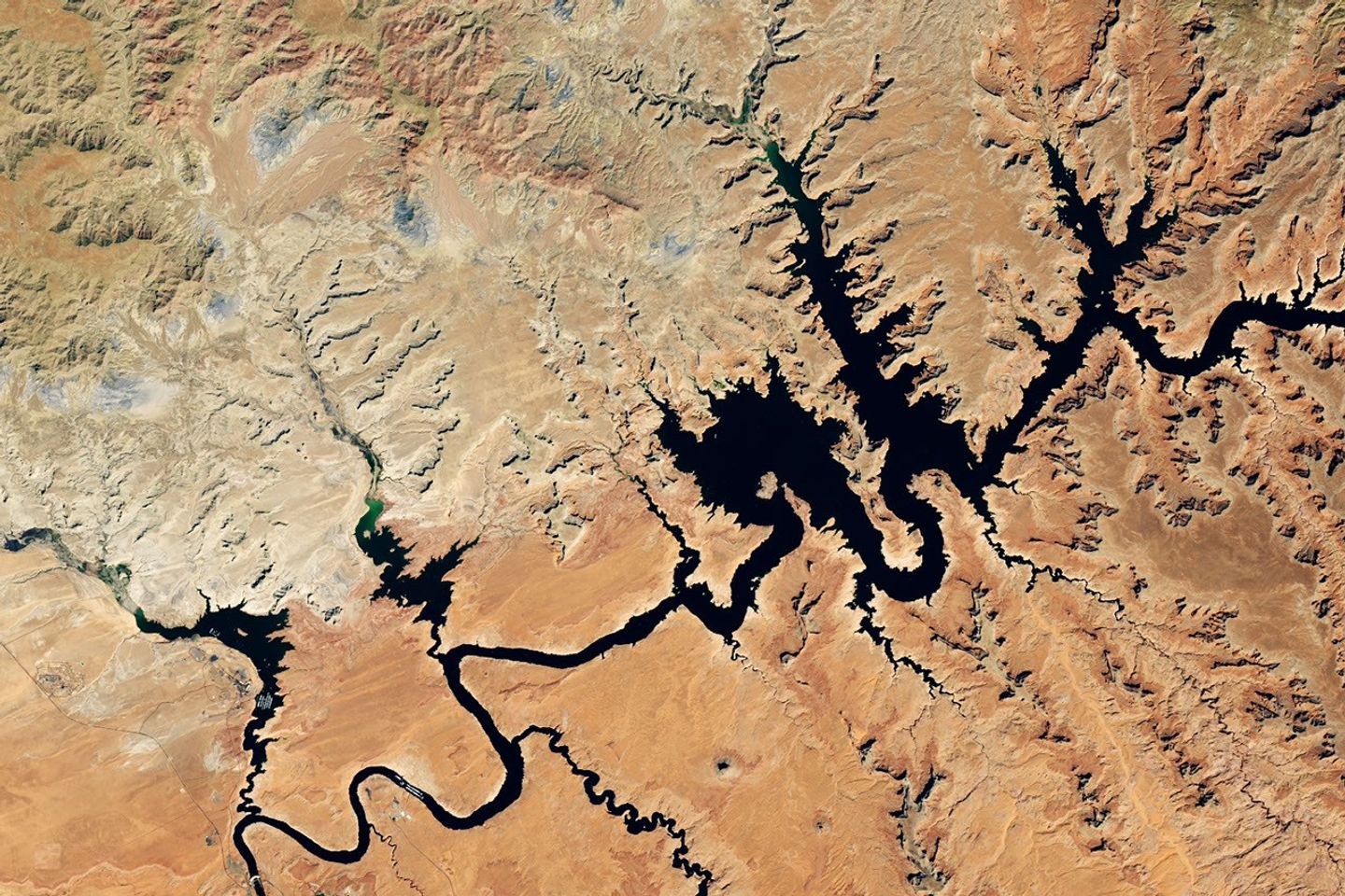 Water in Lake Powell is at record-low levels and looks like a wide river winding through a light orange desert landscape. The Glen Canyon Dam and the town of Page, Arizona, are visible in the lower left.