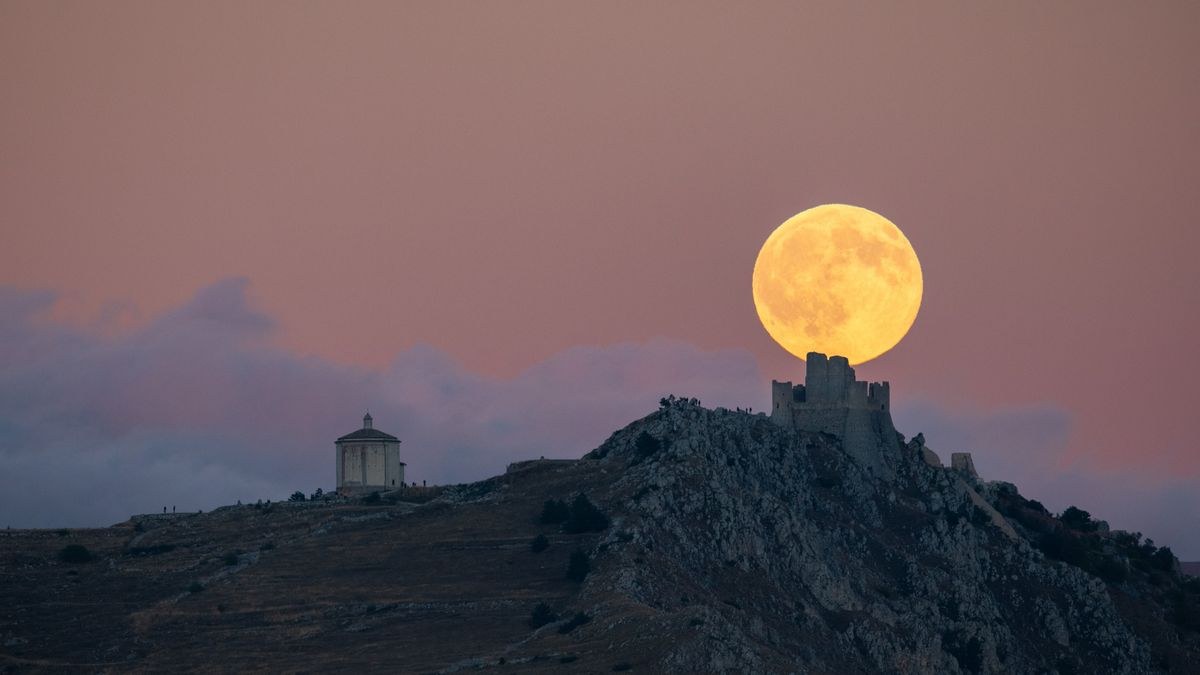 A large golden full moon rises behind the silhouette of Rocca Calascio Castle and the Church of Santa Maria della Piet&amp;agrave; in Calascio, Italy.