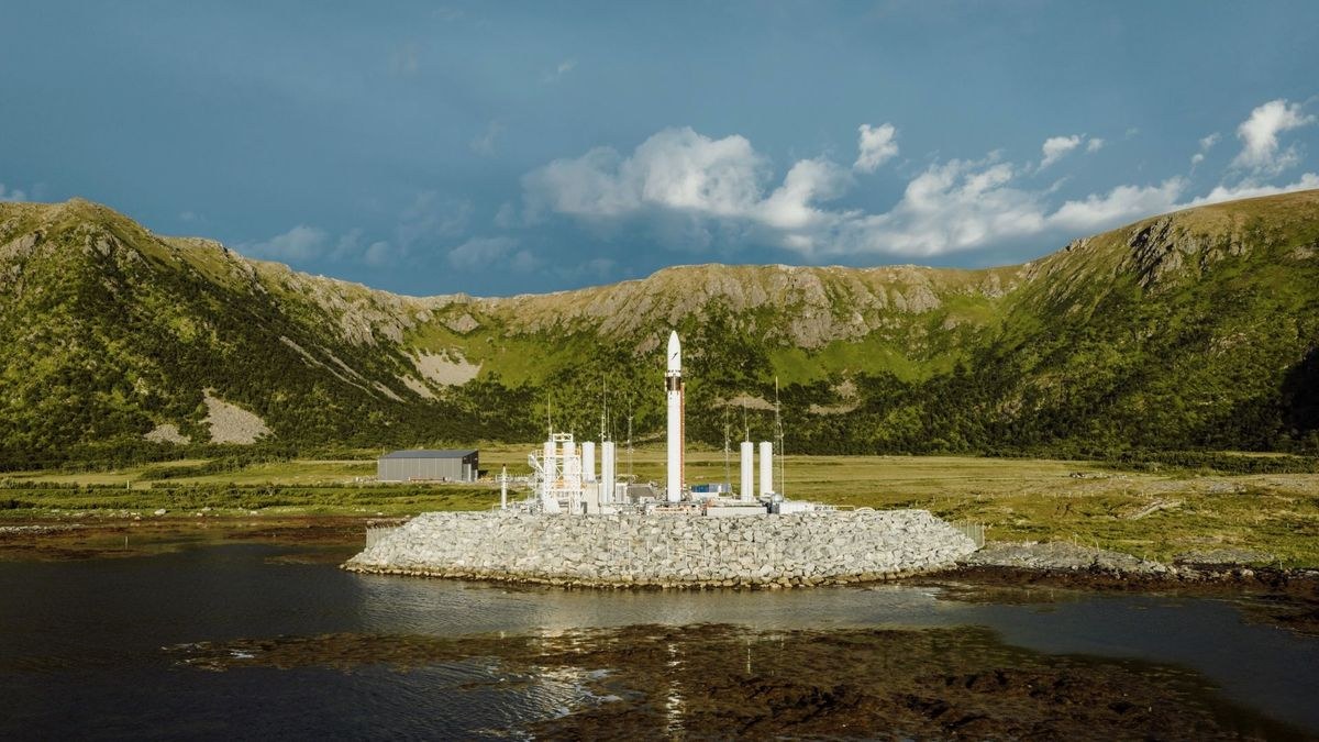 a white rocket stands on a seaside pad with green mountains in the background