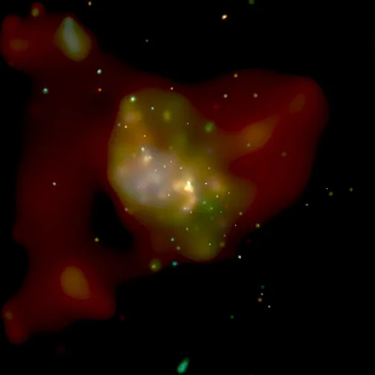 An abstract, brightly glowing cloud shape sits centered against a solid black background. The core of the formation glows bright white and yellow, surrounded by a greenish layer that diffuses into a larger, deep red outer haze. Tiny, multicolored pinpricks of light—mostly yellow, green, and white—are scattered across the central core and the dark space surrounding it.
