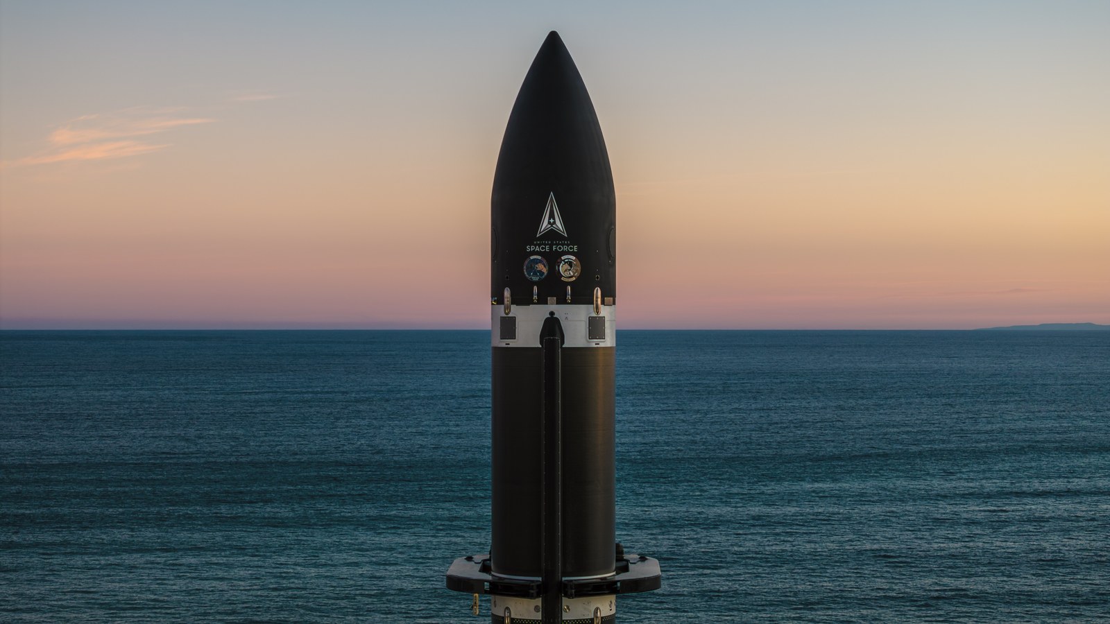 The top of a black rocket stands against a background of rich sea parted in the center by the horizon, and a cool, orange-hued sky. 