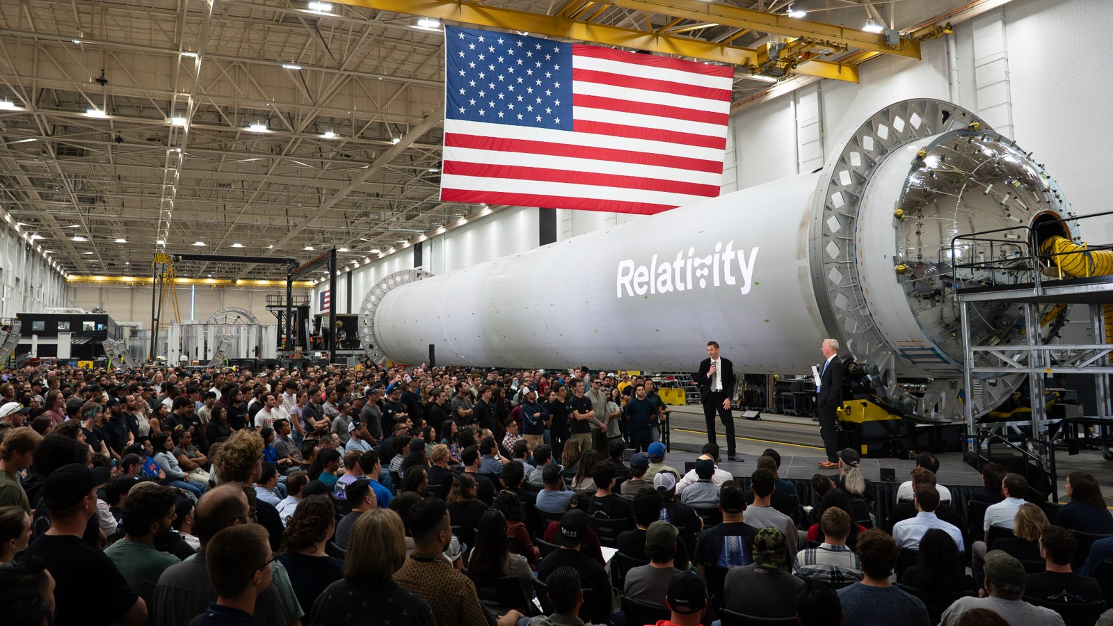 a large crowd sits facing to men in suits at one end of a rocket stage laid horizontally.