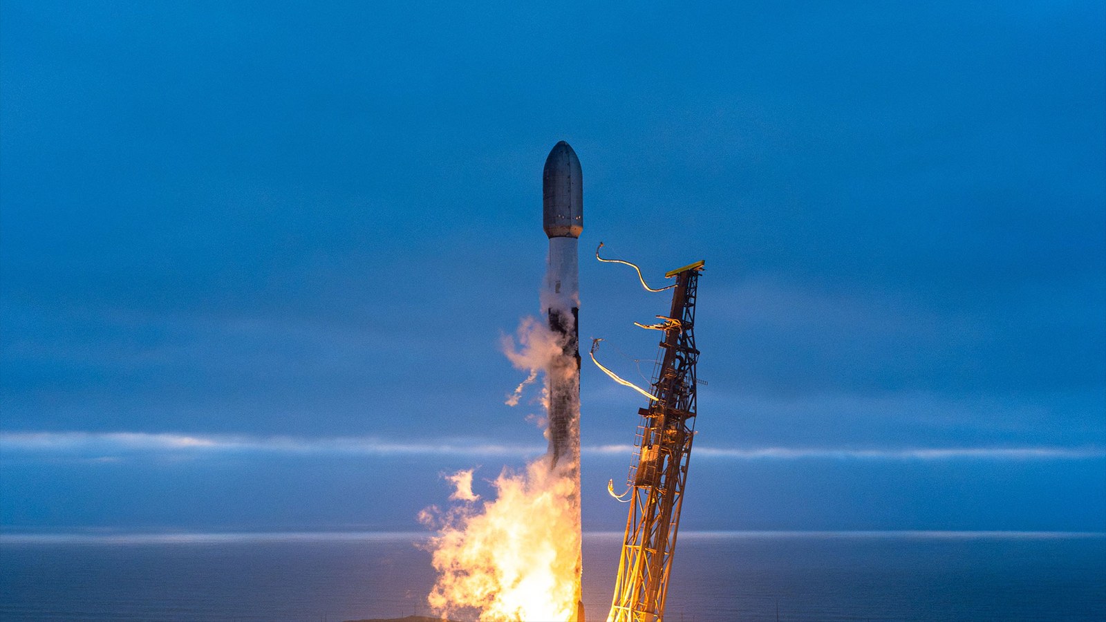 A white and black rocket launches into an overcast evening sky from an ocean-side pad.