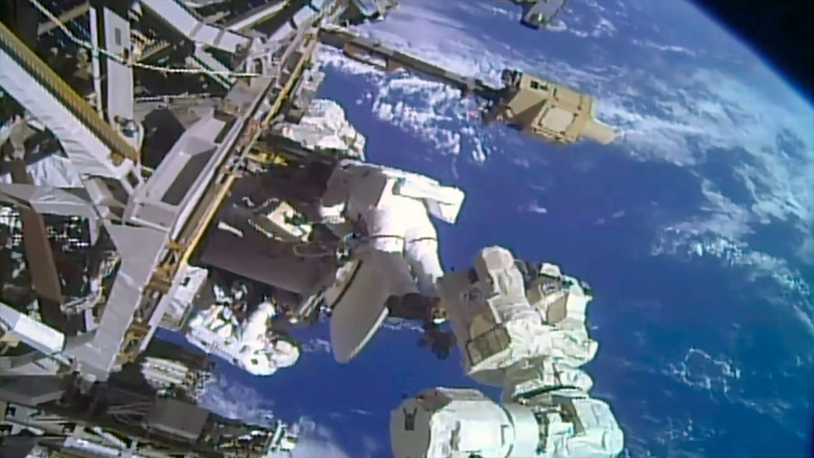two astronauts in white spacesuits work near a large antenna dish outside a space station with a blue and white planet as their backdrop