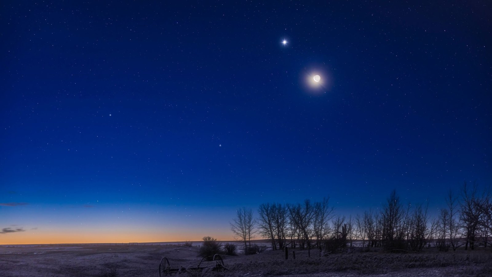 The moon shines in the evening sky with a bright star-like planet glowing close to its upper left above a snowy field lined with trees. The glow of the setting sun can be seen brightening the horizon.