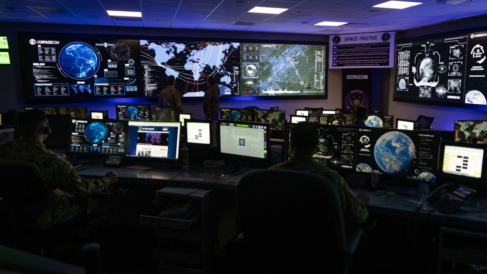 people in military fatigues sit behind computer consoles in a room full of screens showing earth and charts of satellites in orbit