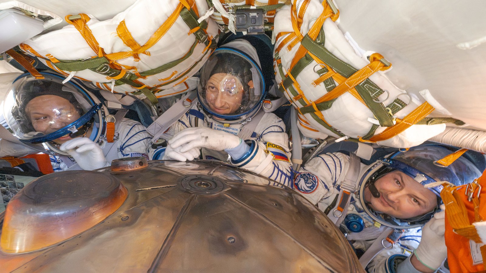 three men in white and blue striped pressure suits are seen from above inside a cramped space capsule