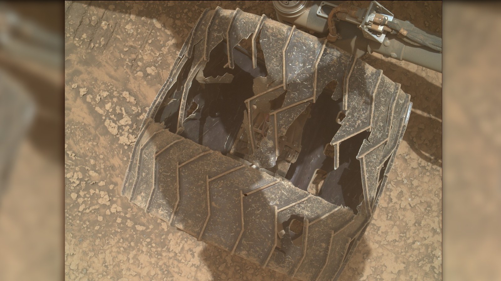 a close-up of a metal wheel with deep cracks and caved-in sections as it rests on a reddish-orange rocky surface