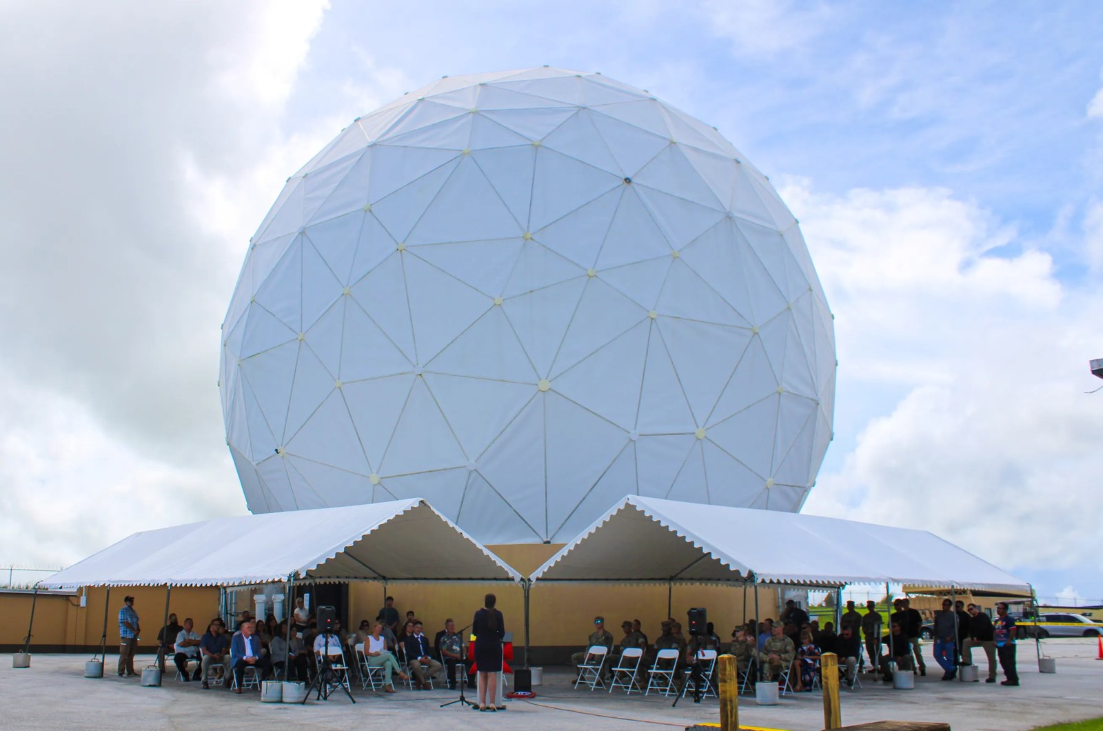 Employees and guests gather beneath white event tents outside a large white geodesic radome during an outdoor ceremony under partly cloudy skies. A woman in a dark suit addresses the audience.
