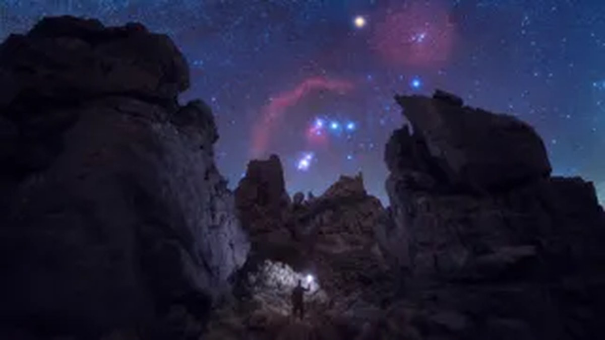 Büyük, pürüzlü kayalar arasında Orion takımyıldızını gösteren bir gökyüzü. Alt kısımda elinde ışık tutan bir insan var.