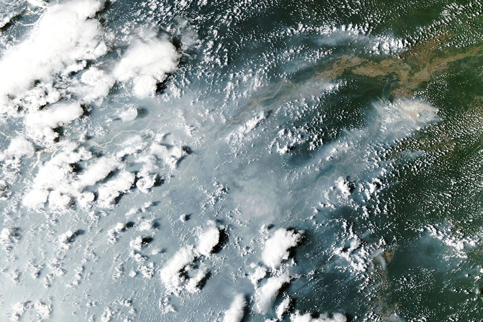 Thick gray smoke partially obscures the Amazon River and surrounding rainforest near the cities of Manaus and Santarém. Puffy white clouds rise above the smoke on the left side of the image.