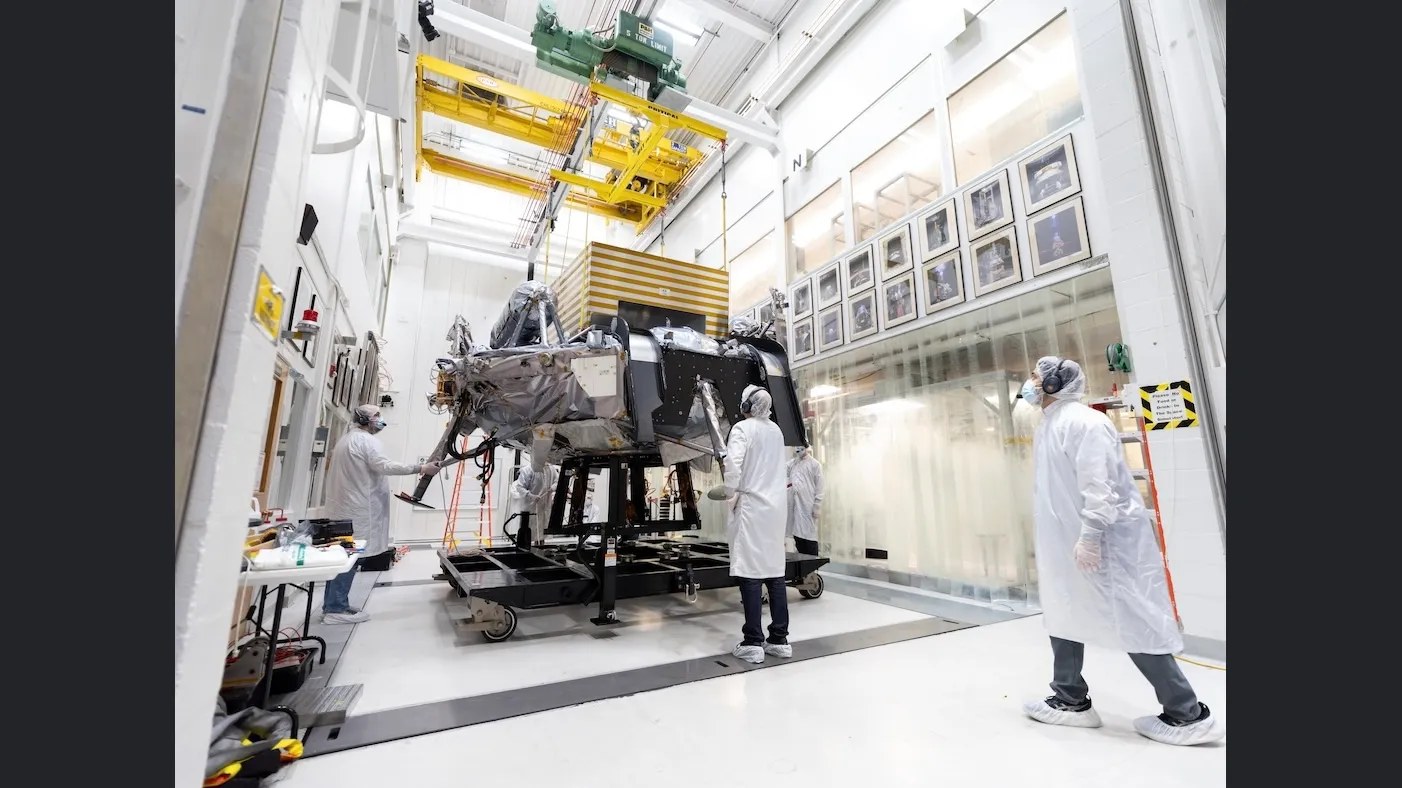 Engineers in white clean-room garb monitor a large spacecraft as it is hoisted by a yellow overhead crane in a hallway in a bright, high-tech laboratory. Framed photos line both sides of the hallway.