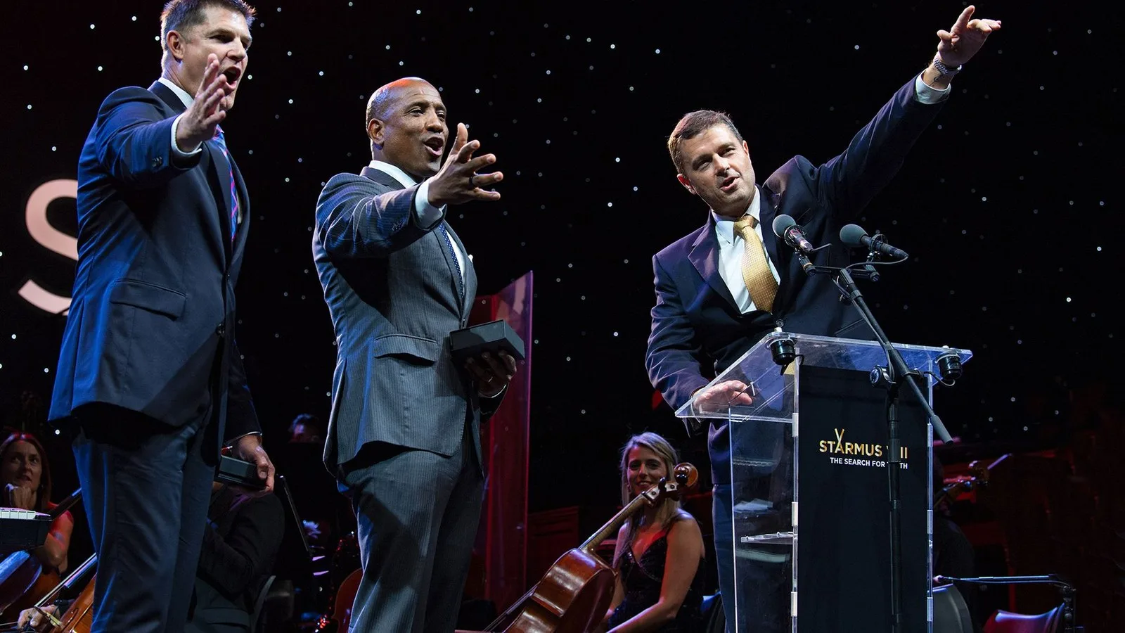 Three men wearing suits stand on a stage and point to an audience.