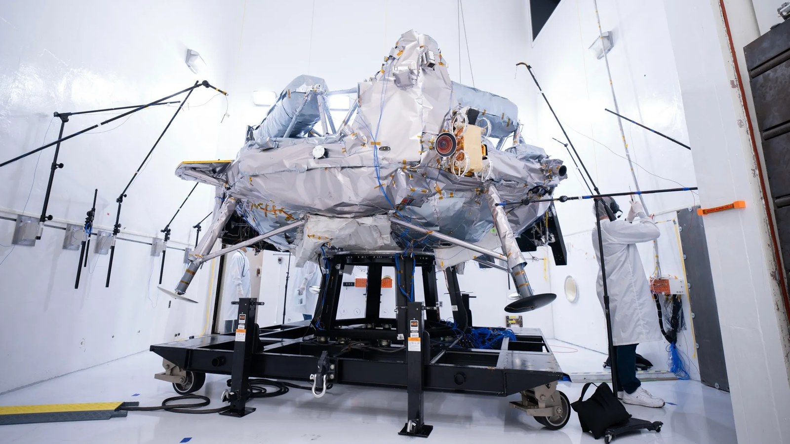 A silvery spacecraft sits on a test frame surrounded by microphones and testing equipment inside a bright white acoustic test chamber, while an engineer in a lab coat works nearby.