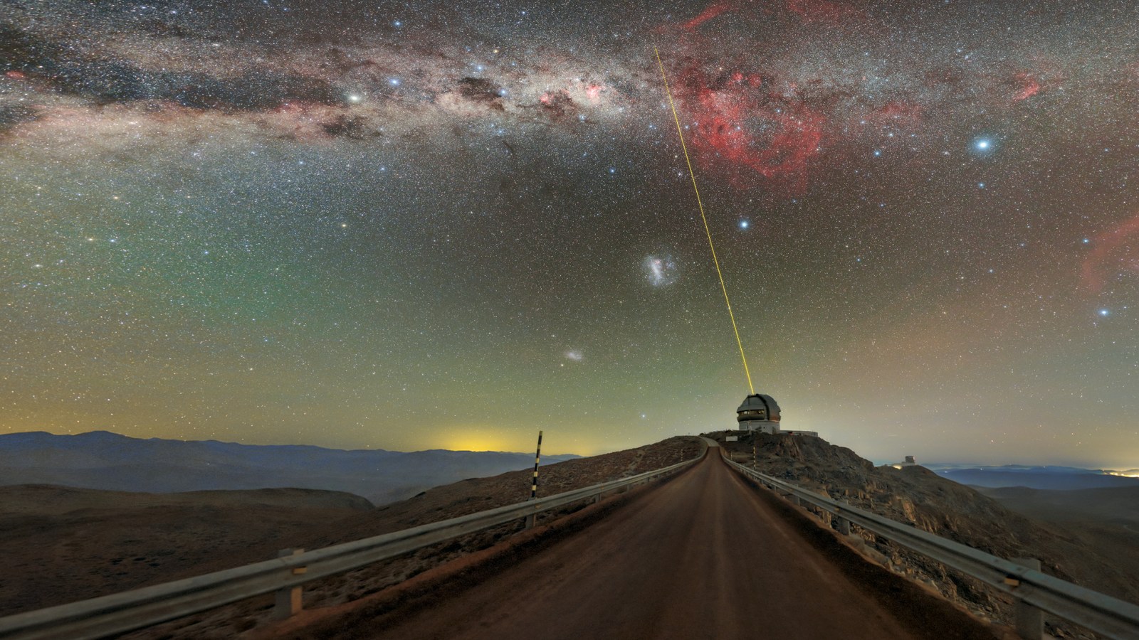 A telescope sits underneath a speckled, colorful night sky as a beam of laser light shoots up as a laser star guide.
